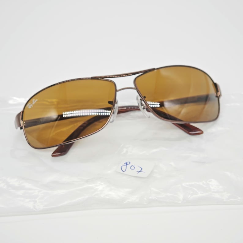 May include: Brown aviator sunglasses with a double bridge and brown tinted lenses. The sunglasses have a metal frame and the Ray-Ban logo is visible on the left lens. The number "807" is written on the plastic bag.