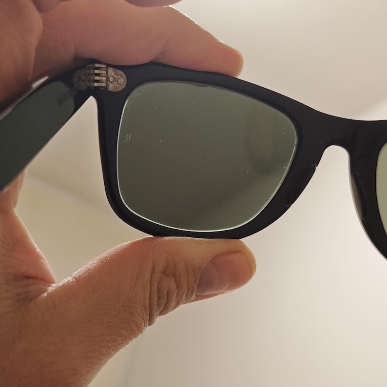 Puede incluir: Gafas de sol negras con un dise&ntilde;o de montura cl&aacute;sico y lentes tintadas en verde oscuro. La montura incluye un mecanismo de bisagra met&aacute;lica visible en la esquina superior, que conecta la patilla con el frente, con un peque&ntilde;o logo grabado.
