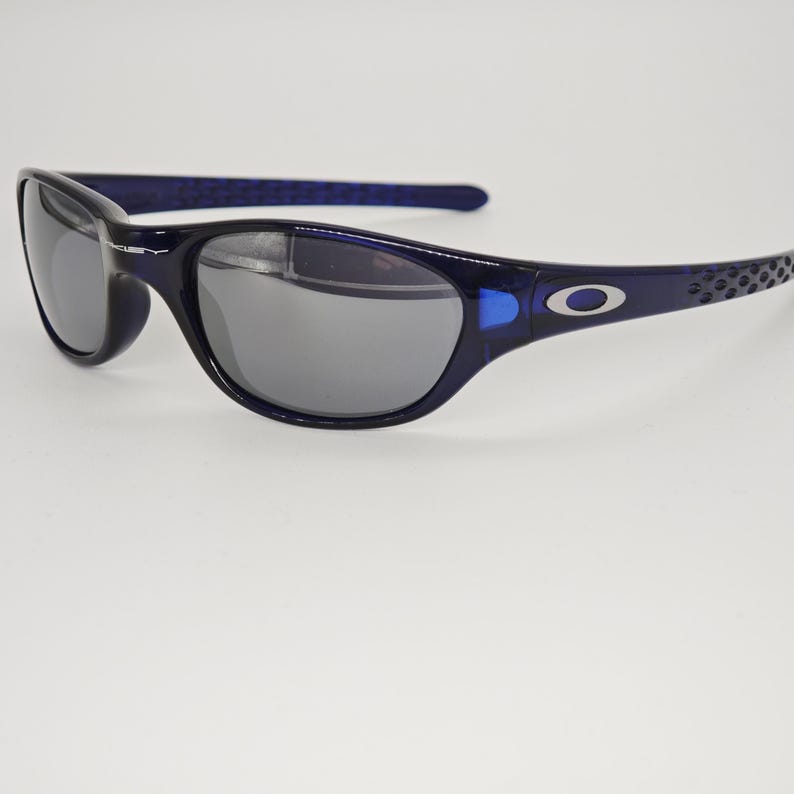May include: Dark blue sunglasses with grey lenses. The sunglasses have a curved frame and a small silver logo on the temples. The arms of the sunglasses have a textured design.