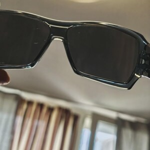 May include: Black rectangular sunglasses with dark lenses. The frames are a glossy black with a subtle textured pattern on the temples. The sunglasses have a modern, sporty design.