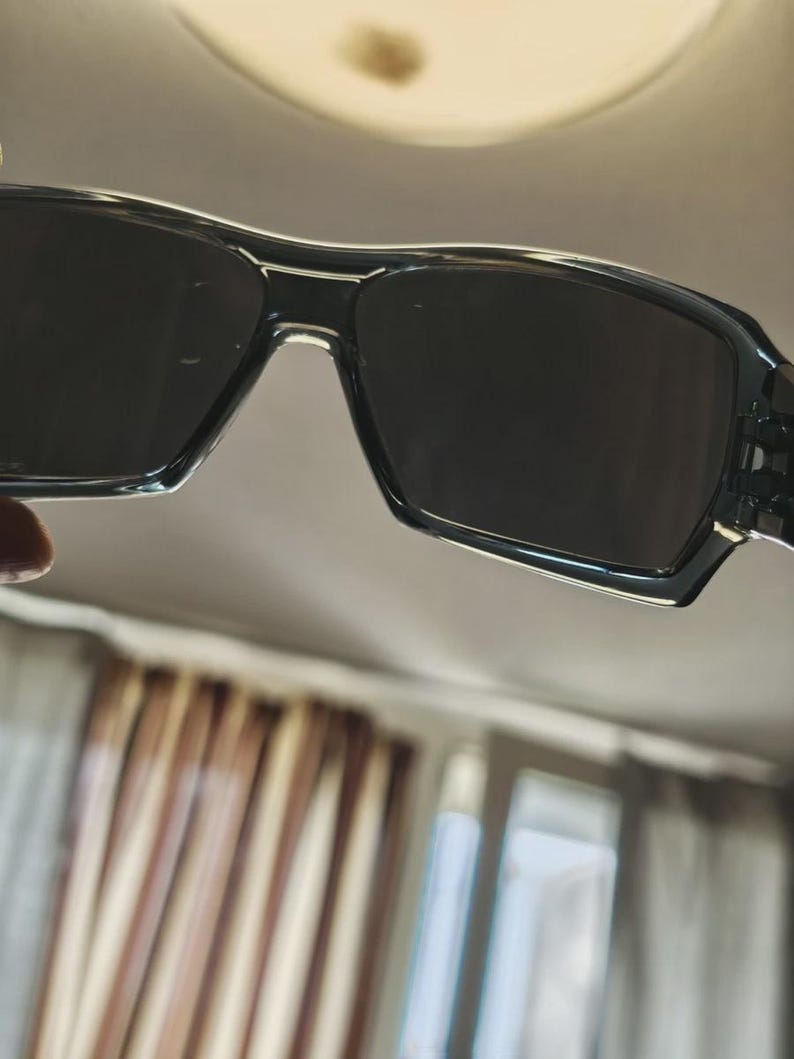 May include: Black rectangular sunglasses with dark lenses. The frames are a glossy black with a subtle textured pattern on the temples. The sunglasses have a modern, sporty design.