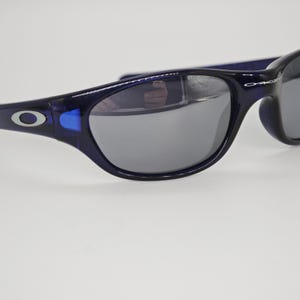 May include: Close-up of dark blue sunglasses with grey lenses. The sunglasses have a curved frame and the brand name "Oakley" is visible on the temple. The lenses reflect the surrounding environment.