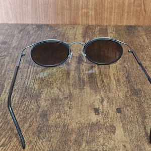 May include: Oval-shaped sunglasses with dark lenses and a thin, black metal frame. The arms of the sunglasses are also black and curve slightly. The sunglasses are set against a wooden background.