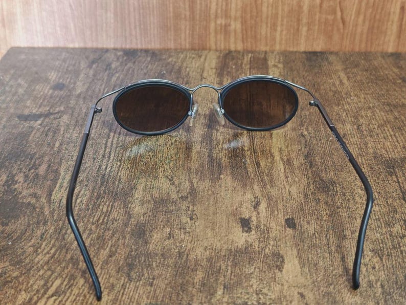 May include: Oval-shaped sunglasses with dark lenses and a thin, black metal frame. The arms of the sunglasses are also black and curve slightly. The sunglasses are set against a wooden background.