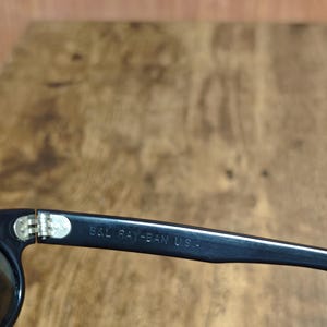 May include: Close-up of black sunglasses with the text "B&L RAY-BAN U.S.A." printed on the arm. The sunglasses have a silver hinge and dark lenses. The frame is a classic, rectangular shape.