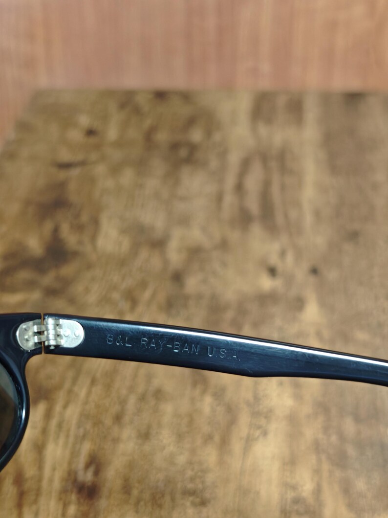 May include: Close-up of black sunglasses with the text "B&L RAY-BAN U.S.A." printed on the arm. The sunglasses have a silver hinge and dark lenses. The frame is a classic, rectangular shape.