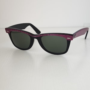 Puede incluir: Gafas de sol con montura negra mate y lentes de color verde oscuro. La parte superior y las patillas presentan un acabado texturizado en color morado intenso. Peque&ntilde;os detalles met&aacute;licos plateados son visibles en las esquinas.