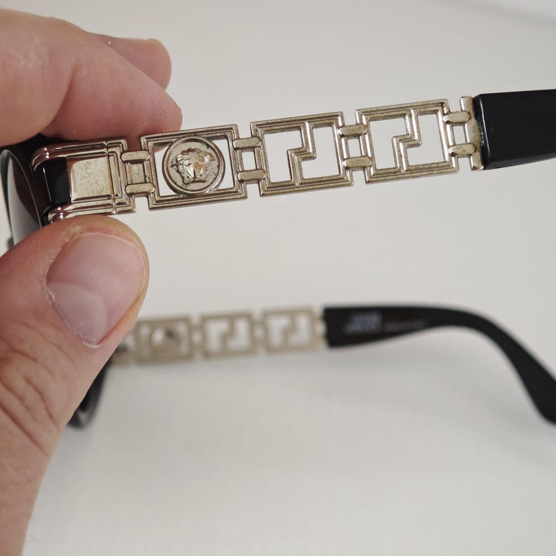 Puede incluir: Gafas de sol negras con varillas de metal plateado que presentan un patr&oacute;n geom&eacute;trico repetitivo y un emblema central de la cabeza de Medusa. Las varillas terminan en extremos de pl&aacute;stico negro s&oacute;lido con un dise&ntilde;o met&aacute;lico detallado.