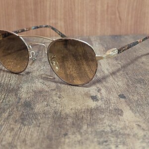 Peut inclure: Lunettes de soleil aviateur &agrave; monture dor&eacute;e avec verres d&eacute;grad&eacute;s marron. Les branches pr&eacute;sentent un motif &agrave; motifs dans les tons marron, bleu et beige. Les lunettes sont pos&eacute;es sur une surface en bois.