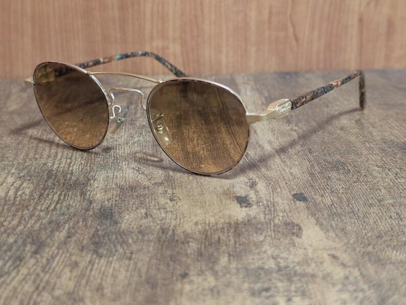 Peut inclure: Lunettes de soleil aviateur &agrave; monture dor&eacute;e avec verres d&eacute;grad&eacute;s marron. Les branches pr&eacute;sentent un motif &agrave; motifs dans les tons marron, bleu et beige. Les lunettes sont pos&eacute;es sur une surface en bois.