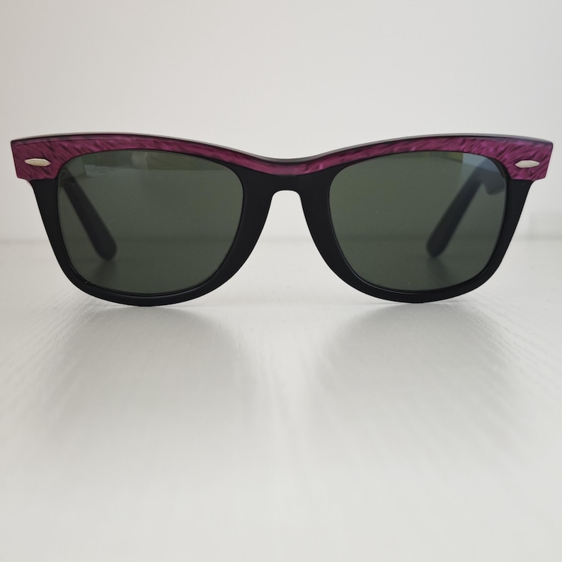 Puede incluir: Gafas de sol con montura negra mate y lentes de color verde oscuro. La parte superior del marco presenta un acabado texturizado en color morado intenso. Peque&ntilde;os detalles met&aacute;licos plateados est&aacute;n incrustados en las esquinas exteriores.