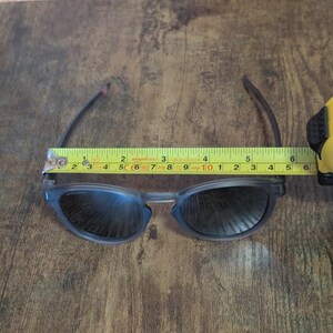 May include: A pair of blue-grey sunglasses with dark lenses, measuring approximately 6 inches in width. The glasses are being measured with a yellow tape measure on a brown wooden surface. The glasses have a modern, sleek design.