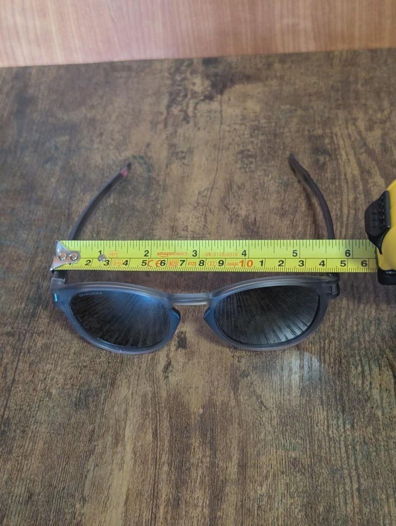 May include: A pair of blue-grey sunglasses with dark lenses, measuring approximately 6 inches in width. The glasses are being measured with a yellow tape measure on a brown wooden surface. The glasses have a modern, sleek design.