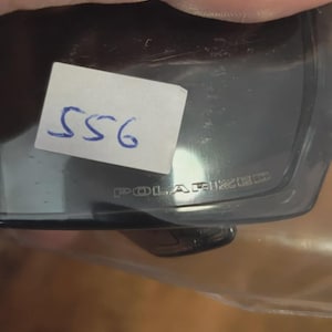 May include: Dark gray polarized sunglasses with the word "POLARIZED" printed on the frame. A small white sticker with the number "556" written in blue ink is attached to the sunglasses.