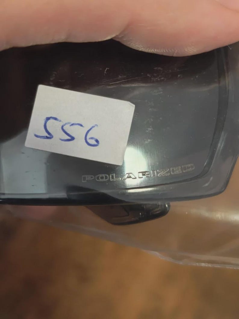 May include: Dark gray polarized sunglasses with the word "POLARIZED" printed on the frame. A small white sticker with the number "556" written in blue ink is attached to the sunglasses.