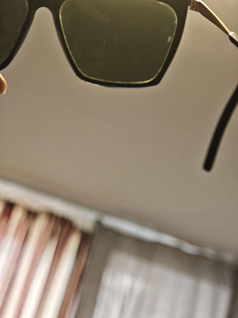 Peut inclure: Lunettes de soleil noires avec une monture dor&eacute;e et des verres vert fonc&eacute;. Les lunettes de soleil ont une forme carr&eacute;e et sont pr&eacute;sent&eacute;es sur un fond neutre. Les branches des lunettes sont noires.