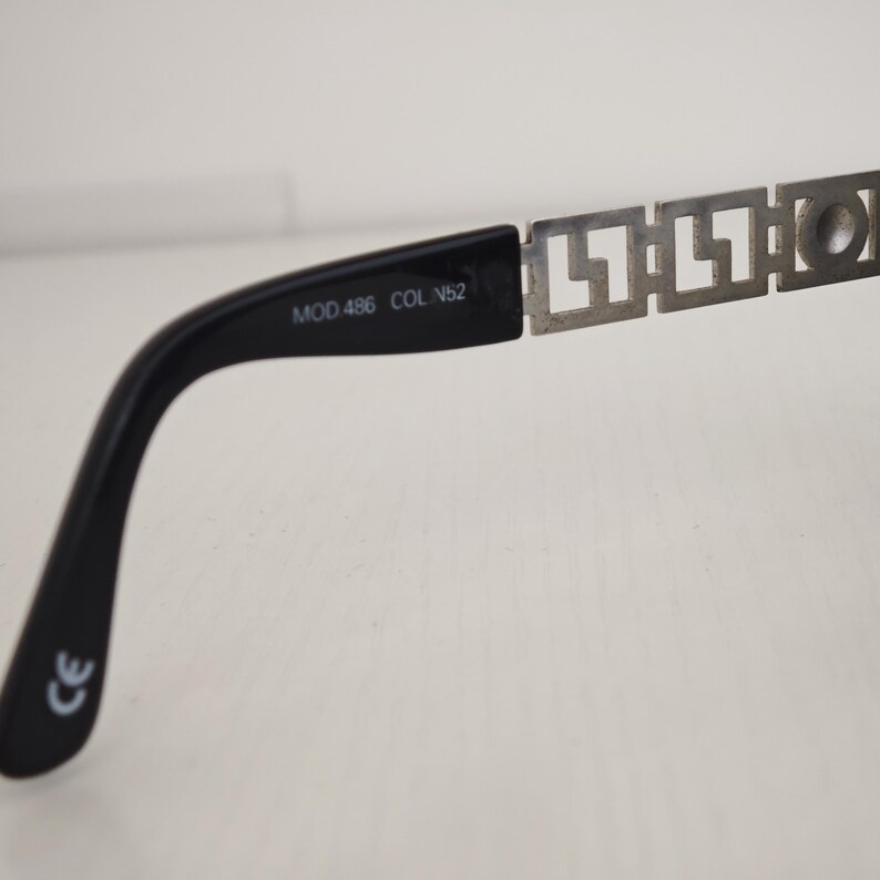 Puede incluir: Una patilla negra de gafas con una bisagra de metal plateado con un patr&oacute;n geom&eacute;trico de greca. El interior de la patilla de pl&aacute;stico negro tiene impreso el texto MOD.486 COL.N52 y una marca CE cerca del extremo curvado.