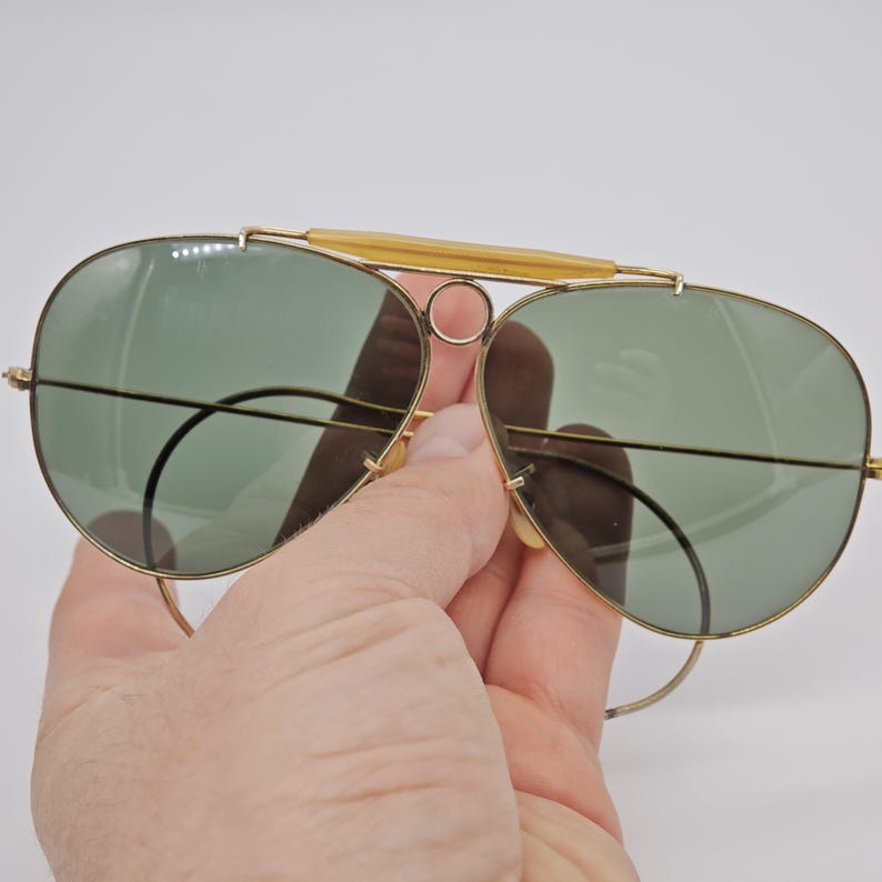 May include: Gold-framed aviator sunglasses with green lenses. The sunglasses have a double bridge and a bar across the top. The lenses are teardrop-shaped and the frames are thin and delicate.