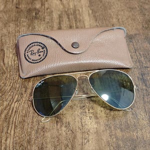 May include: Gold-framed aviator sunglasses with dark lenses and a tan leather case. The case has a button closure and a Ray-Ban logo. The sunglasses are on a wooden surface.