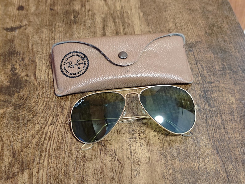 May include: Gold-framed aviator sunglasses with dark lenses and a tan leather case. The case has a button closure and a Ray-Ban logo. The sunglasses are on a wooden surface.