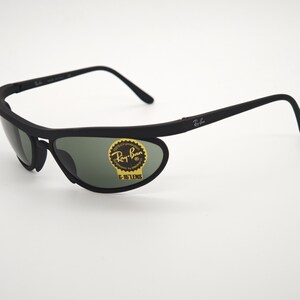 May include: Black Ray-Ban sunglasses with green lenses. The sunglasses have a unique wraparound design and a sticker on the lens that reads "Ray-Ban G-15 LENS". The frames are matte black.