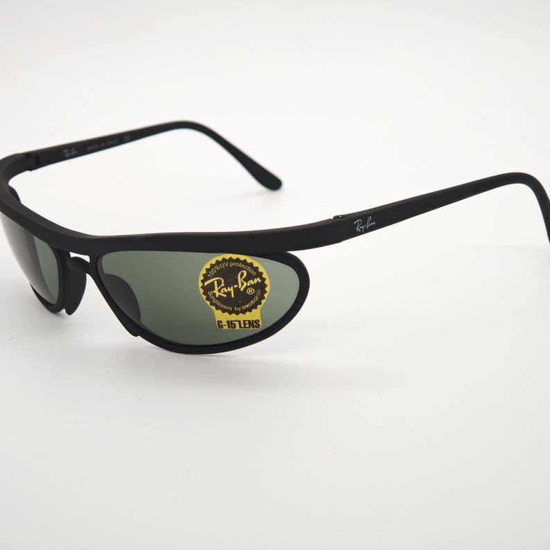 May include: Black Ray-Ban sunglasses with green lenses. The sunglasses have a unique wraparound design and a sticker on the lens that reads "Ray-Ban G-15 LENS". The frames are matte black.