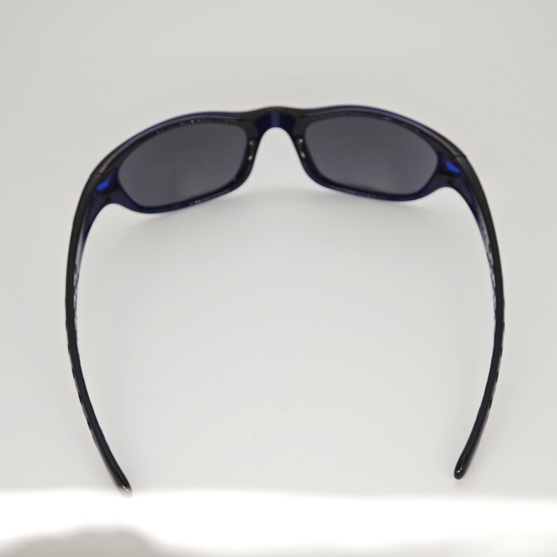 May include: Dark blue sunglasses with dark grey lenses. The sunglasses have a curved frame and temples, and are set against a white background. The sunglasses are designed for outdoor use.