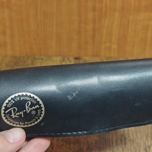 May include: Black Ray-Ban sunglasses case with a gold and white logo. The case is made of a textured material and has a rounded shape. The logo reads "Ray-Ban Sunglasses by Bausch & Lomb" with "100% UV protection" text.