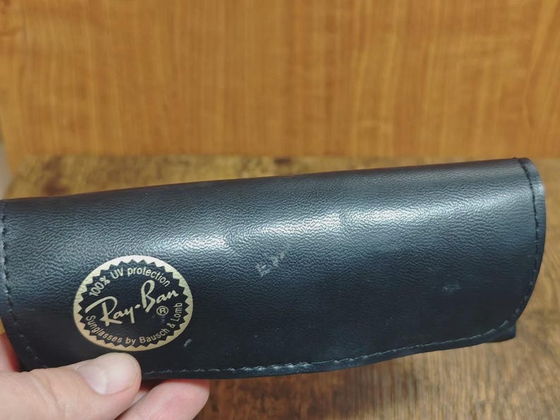 May include: Black Ray-Ban sunglasses case with a gold and white logo. The case is made of a textured material and has a rounded shape. The logo reads "Ray-Ban Sunglasses by Bausch & Lomb" with "100% UV protection" text.