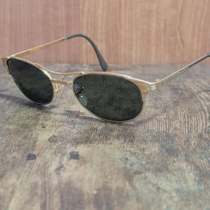 May include: Gold-tone sunglasses with dark lenses. The frames have a thin, oval shape with a double bridge and black earpieces. The sunglasses are set against a wooden background.