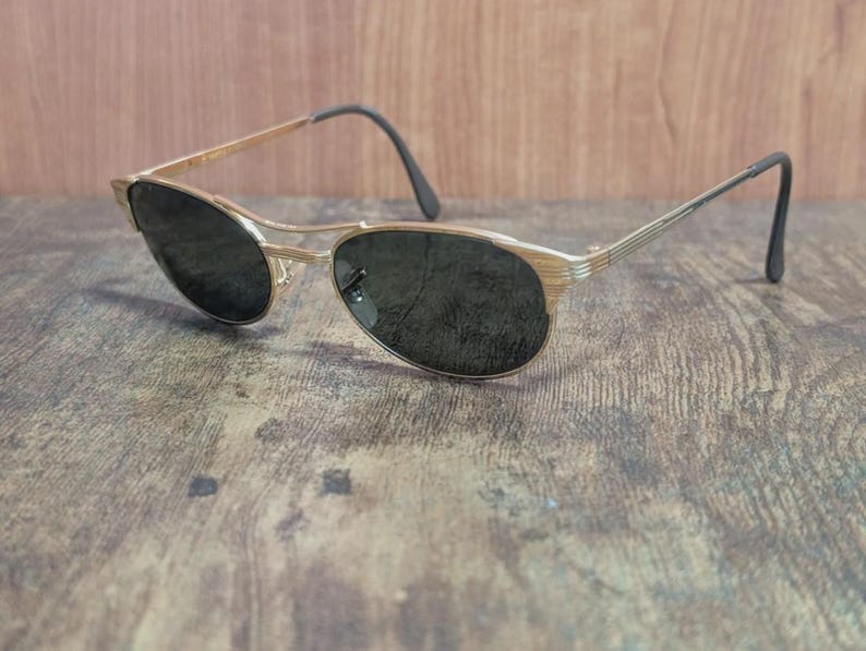 May include: Gold-tone sunglasses with dark lenses. The frames have a thin, oval shape with a double bridge and black earpieces. The sunglasses are set against a wooden background.