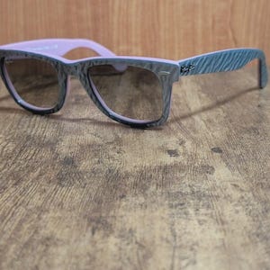 Puede incluir: Gafas de sol grises y rosas con un patr&oacute;n texturizado en las monturas y las patillas. Las lentes son de color marr&oacute;n oscuro. Las gafas de sol est&aacute;n sobre una superficie de madera.