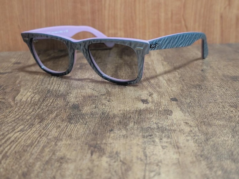 Puede incluir: Gafas de sol grises y rosas con un patr&oacute;n texturizado en las monturas y las patillas. Las lentes son de color marr&oacute;n oscuro. Las gafas de sol est&aacute;n sobre una superficie de madera.