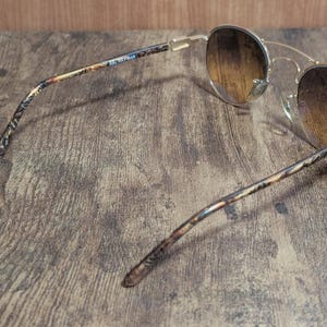 Peut inclure: Lunettes de soleil dor&eacute;es avec des verres d&eacute;grad&eacute;s marron. Les branches pr&eacute;sentent un motif avec le texte "BEL RAFRAN". Les lunettes de soleil sont pos&eacute;es sur un fond en bois.