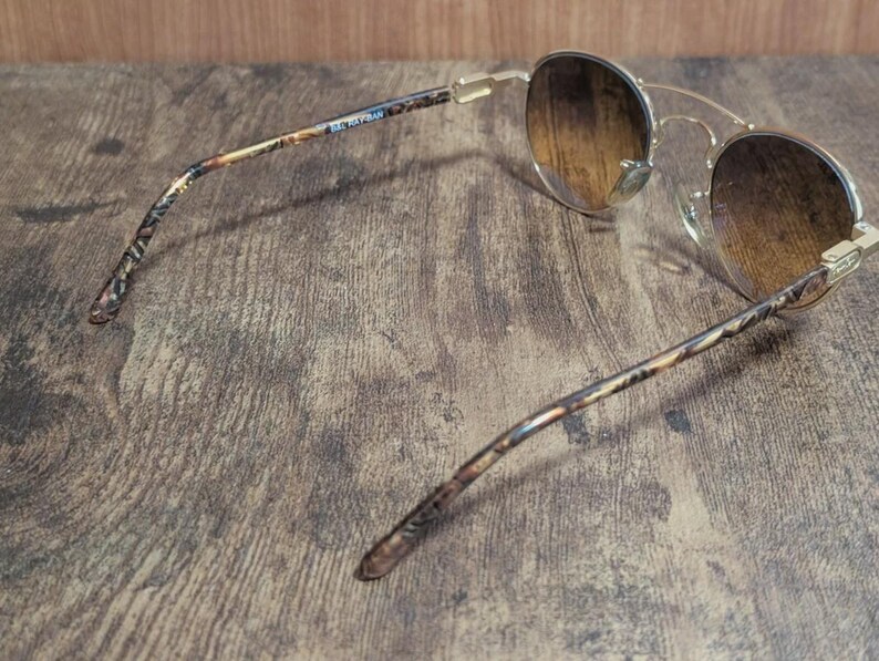 Peut inclure: Lunettes de soleil dor&eacute;es avec des verres d&eacute;grad&eacute;s marron. Les branches pr&eacute;sentent un motif avec le texte "BEL RAFRAN". Les lunettes de soleil sont pos&eacute;es sur un fond en bois.