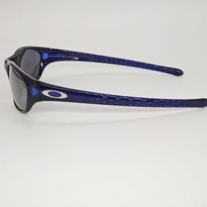 May include: Dark blue sunglasses with black lenses. The arms feature a textured design. The Oakley logo is visible on the side. These sunglasses are suitable for sports and outdoor use. The frame is a dark navy blue.