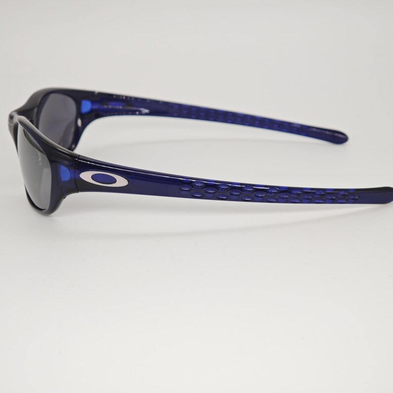 May include: Dark blue sunglasses with black lenses. The arms feature a textured design. The Oakley logo is visible on the side. These sunglasses are suitable for sports and outdoor use. The frame is a dark navy blue.