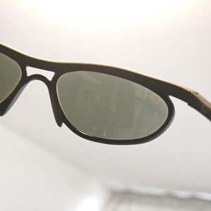 May include: Close-up of black sunglasses with oval lenses and a dark frame. The lenses are a muted green colour, and the frame has a matte finish. The sunglasses are designed for sports or outdoor activities.
