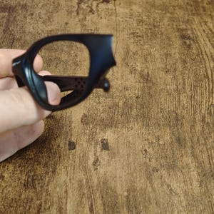 Op de afbeelding: Zwart brillenmontuur met een afgeronde rechthoekige vorm. Het montuur wordt in een hand gehouden en is gemaakt van een mat materiaal. Het montuur heeft een kleine neusbrug en veren. Het montuur staat tegen een houten achtergrond.
