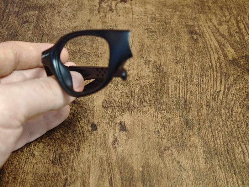 Op de afbeelding: Zwart brillenmontuur met een afgeronde rechthoekige vorm. Het montuur wordt in een hand gehouden en is gemaakt van een mat materiaal. Het montuur heeft een kleine neusbrug en veren. Het montuur staat tegen een houten achtergrond.