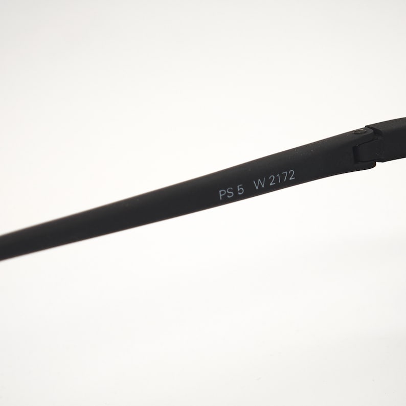 May include: A close-up of a black eyeglass frame arm with the text "PS 5 W 2172" printed in white. The arm is angled and appears to be made of a matte material against a white background.