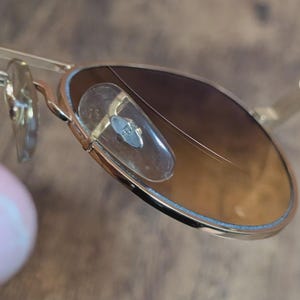 Peut inclure: Gros plan de lunettes de soleil &agrave; monture dor&eacute;e avec verres d&eacute;grad&eacute;s marron. Les lunettes ont un style aviateur classique avec un double pont et des plaquettes nasales transparentes. Les verres pr&eacute;sentent une rayure.