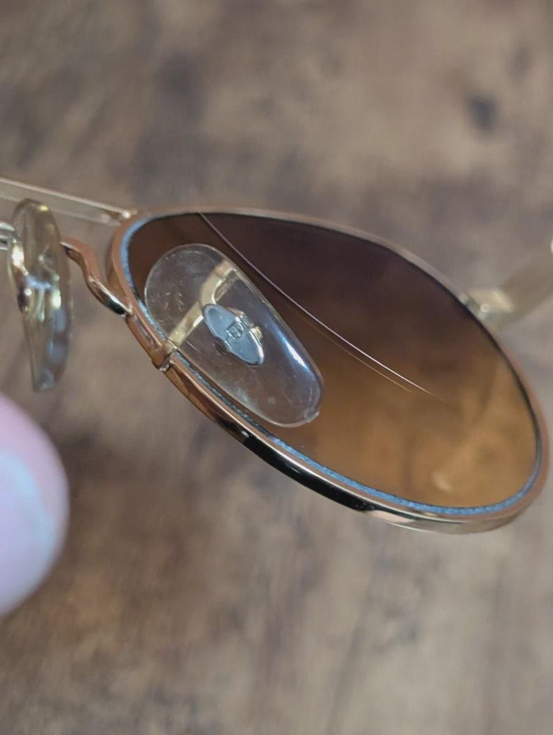 Peut inclure: Gros plan de lunettes de soleil &agrave; monture dor&eacute;e avec verres d&eacute;grad&eacute;s marron. Les lunettes ont un style aviateur classique avec un double pont et des plaquettes nasales transparentes. Les verres pr&eacute;sentent une rayure.