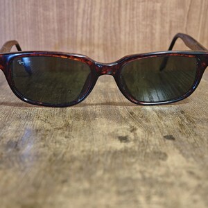 May include: Tortoiseshell sunglasses with dark lenses. The frames have a classic, rectangular shape with a subtle curve at the top. The Ray-Ban logo is visible on the left lens. The sunglasses are set against a wooden background.