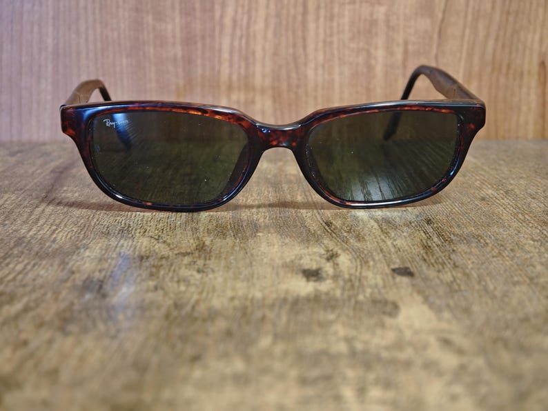 May include: Tortoiseshell sunglasses with dark lenses. The frames have a classic, rectangular shape with a subtle curve at the top. The Ray-Ban logo is visible on the left lens. The sunglasses are set against a wooden background.
