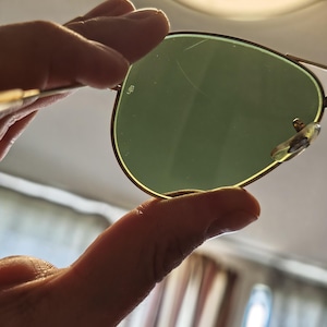 May include: Aviator-style sunglasses with a gold-colored frame and green lenses. The lenses have a slight scratch. The sunglasses are held up against a bright background.
