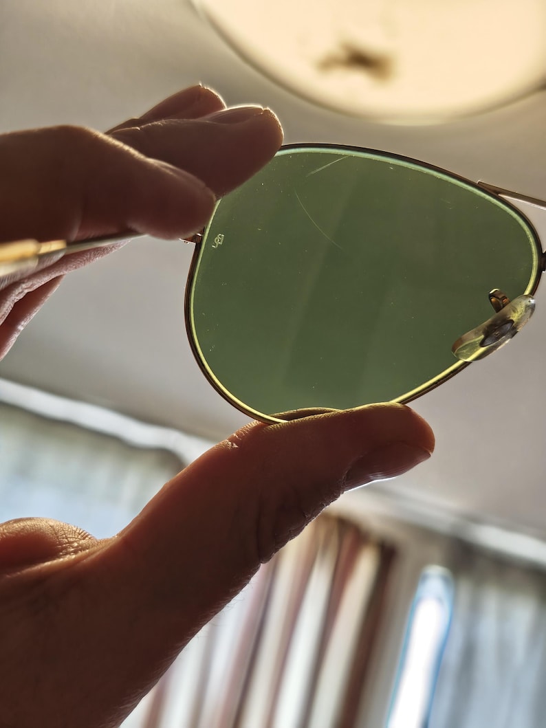 May include: Aviator-style sunglasses with a gold-colored frame and green lenses. The lenses have a slight scratch. The sunglasses are held up against a bright background.