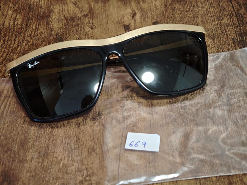 Peut inclure: Lunettes de soleil noires avec une monture sup&eacute;rieure dor&eacute;e. Les lunettes ont des verres fonc&eacute;s et une forme rectangulaire. Le logo Ray-Ban est visible sur la lentille gauche. Une petite &eacute;tiquette blanche avec le num&eacute;ro 669 est &eacute;galement pr&eacute;sente.
