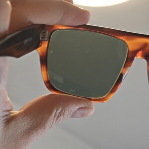 May include: Close-up of sunglasses with a tortoiseshell frame and green lenses. The rectangular lenses are set in a thick, patterned frame. The sunglasses are held up, showing the hinge and the inside of the frame.