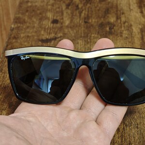 Peut inclure: Lunettes de soleil noires avec une monture sup&eacute;rieure dor&eacute;e. Les lunettes ont des verres fonc&eacute;s et le logo Ray-Ban est visible sur la lentille gauche. Les lunettes sont tenues dans une main.